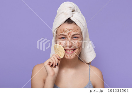 Young happy woman with moisturizing facial mask, holds sponge for removing makeup, has beauty treatments, has fresh skin after taking shower, poses against lilac background. 101331502