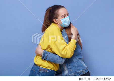 Horizontal image of two young women being infected, wearing protection masks, hugging each other, being in quarantine, breaking coronavirus safety measures. People and coronavirus spread concept. 101331602