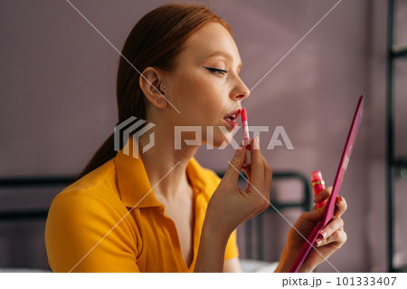 Close-up side view of pretty young woman applying lipstick, holding mirror and looking at herself. Portrait of beauty redhead female looking facial at mirror for makeup cosmetic with lips elegance. 101333407