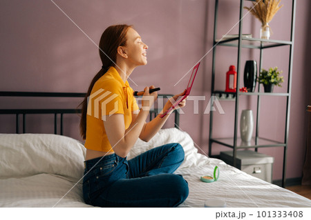 Side view of cheerful redhead young woman applying nude facial powder to get natural effect with big professional makeup brush, looking in mirror. Concept steps of make-up applying yourself. Side view of cheerful redhead young woman applying nude facial powder to get natural effect with big professional makeup brush, looking in mirror. Concept steps of make-up applying yourself. 101333408