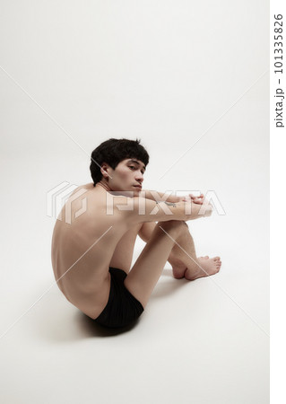 Studio portrait of young man posing shirtless, sitting on floor against white background. 101335826