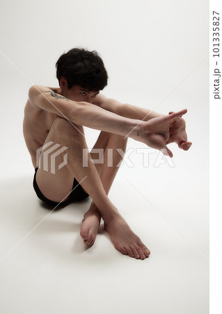 Portrait of young man posing shirtless, sitting, hiding head in hands on floor against white studio background. 101335827