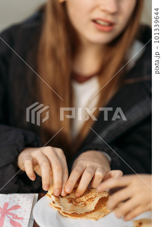 Young girl's hands in black jacket holding crepes in cafe outdoors while she is talking to her friend Young girl's hands in black jacket holding crepes in cafe outdoors while she is talking to her friend 101339644