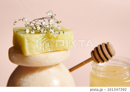 Selective focus on a bar of cold-pressed homemade natural organic soap with floral and herbal ingredients, displayed on a beige marble surface against a jar with honey, on pink pastel background 101347392