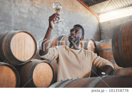 Wine expert or entrepreneur with glass during tasting test in a cellar or distillery in a warehouse. Black agriculture worker in alcohol production storage or wooden barrel in a manufacturing winery 101358776