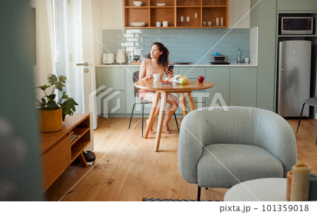 Woman on phone thinking of breakfast idea, healthy snack while drinking coffee in the morning in stylish kitchen. Relaxed and alone girl looking nostalgic in a modern apartment with interior design 101359018