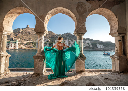 Rear view of a happy blonde woman in a long mint dress posing against the backdrop of the sea in an old building with columns. Girl in nature against the blue sky. 101359358