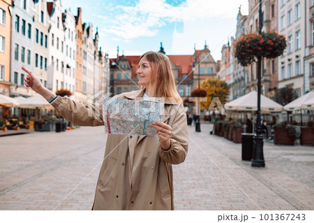 Happy woman tourist pointing finger and holding a paper map, searching direction on location map during vacation in Gdansk old town. Happy woman tourist pointing finger and holding a paper map, searching direction on location map during vacation in Gdansk old town. 101367243