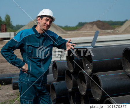 Caucasian male builder in a hard hat stands near the pipes and uses a laptop at a construction site.  101370875