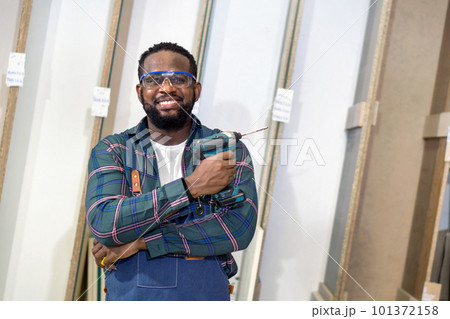 Short black hair man with moustache and beard stand with arm folded, holding cordless electric drill. Plywood storage compartment are on the background. Carpenter worker work in a furniture factory. Short black hair man with moustache and beard stand with arm folded, holding cordless electric drill. Plywood storage compartment are on the background. Carpenter worker work in a furniture factory. 101372158