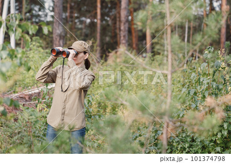 A woman forester in uniform looks through binoculars and monitoring the forest area in summer, selective focus. 101374798