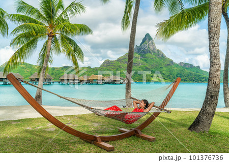 Luxury resort beach holiday woman relaxing lying down in hammock by the overwater bungalows of hotel in Bora Bora, Tahiti, French Polynesia. Happy summer vacation getaway. Luxury resort beach holiday woman relaxing lying down in hammock by the overwater bungalows of hotel in Bora Bora, Tahiti, French Polynesia. Happy summer vacation getaway. 101376736