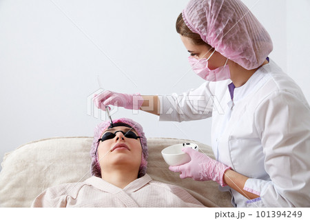 Carbon peeling in beauty salon. Cosmetologist applying black mask on the face of a beautiful woman for carbon peeling 101394249