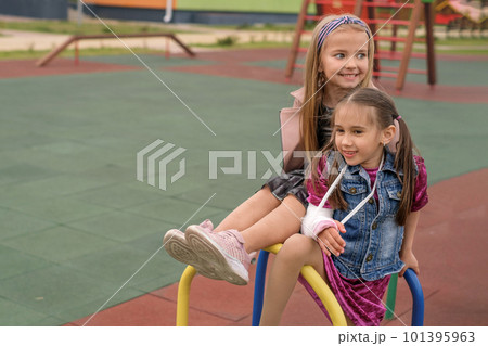 Girl with fractures hand and her friend are sitting on the playground crossbar at the kindergarten. Girl with fractures hand and her friend are sitting on the playground crossbar at the kindergarten. 101395963