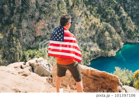Young man standing on a rock cliff with US flag on his back and looking at sea beneath. Veteran traveller wearing American flag on his back while taking mountain hike. 4 fourth July Independence Day. 101397106