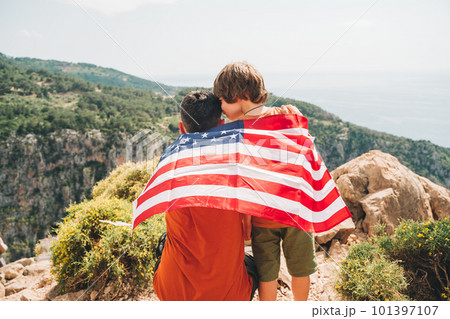 Young man dad father and his son school boy kid standing on a rock cliff with US flag on shoulders and looking at sea. Travellers child wearing American flag on back. 4 July Independence Day. 101397107