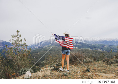 Young woman standing on a rock cliff and waving the US flag while looking at mountains in the background. Girl traveller waving American flag standing on mountain top. 4 fourth July Independence Day 101397108