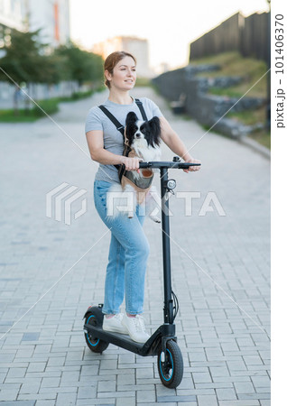 A woman rides an electric scooter with a dog in a backpack. Pappilion Spaniel Continental in a sling. Vertical. 101406370
