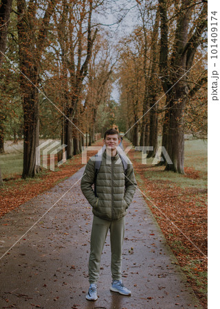 a guy in a jacket stands on an alley in the park during the fall season. Portrait oh handsome teen guy, young man in hoodie, down jacket standing, walking in beautiful golden autumn park, looking at 101409174