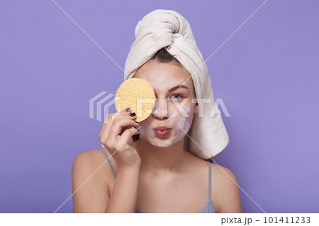 Horizontal shot of young attractive girl posing with white towel on head, holding sponge in hand and covering her eye with it, keeping lips rounded, making kiss gesture, morning cosmetic procedures. 101411233
