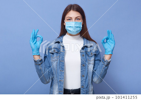 Picture of young woman showing sign ok, invoking to not panic, lady wearing protective face mask and disposable medical gloves, preventing virus infection, isolated over blue background. Coronavirus Picture of young woman showing sign ok, invoking to not panic, lady wearing protective face mask and disposable medical gloves, preventing virus infection, isolated over blue background. Coronavirus 101411255