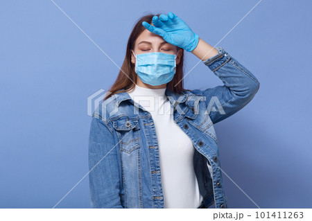 Image of exhausted sick young female having fever, putting one hand on forehead, feeling unwell, closing both eyes, having Covid19, wearing antibacterial mask and gloves. Coronavirus concept. 101411263
