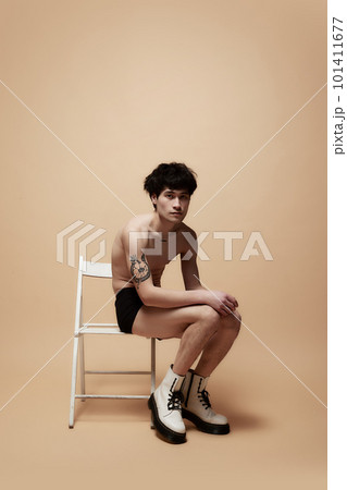 Shy look. Young asian guy in underwear and stylish boots, posing shirtless on chair against light brown studio background 101411677