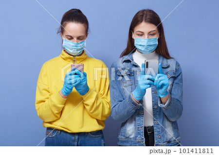 Indoor shot of attentive concentrated two women holding their smartphones, reading news about coronavirus, wearing antibacterial masks and gloves, being in quarantine. Self-isolation concept. 101411811