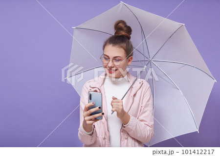Portrait of good looking beautiful young girl holding umbrella and smartphone, wearing accessories, making selfie, shooting video, using social networking sites, having bunch on head. Youth concept. 101412217