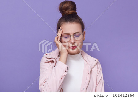 Indoor shot of young woman having headache, putting hand on temple, keeping eyes closed, standing isolated over lilac background, girl wearing casual clothing. Concept of problems, fever, health care. 101412264
