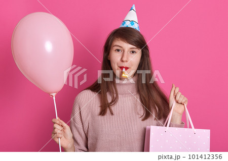 Portrait of beautiful young woman with party whistle and birthday hat, dresses pink sweater, model posing over pastel pink background, holding air baloons. Birthday, holiday party, people concept. Portrait of beautiful young woman with party whistle and birthday hat, dresses pink sweater, model posing over pastel pink background, holding air baloons. Birthday, holiday party, people concept. 101412356