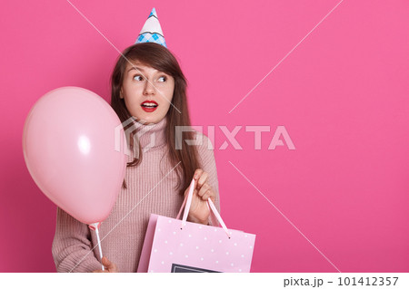 Happy brunette girl with pink balloon and bithday present in hands, keeps mouth opened, being in surprise, looks aside with astinished facial expression, copy space for advertisment or promotion. 101412357