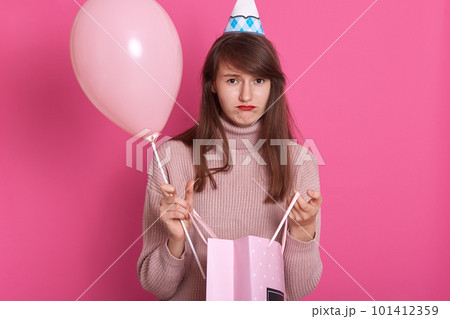 Closeup portrait of beautiful disappointed girl with gift package and balloon, wearing pink sweater and birthday hat, posing isolated over rosy background with pouty lips, having unhappy expression. 101412359