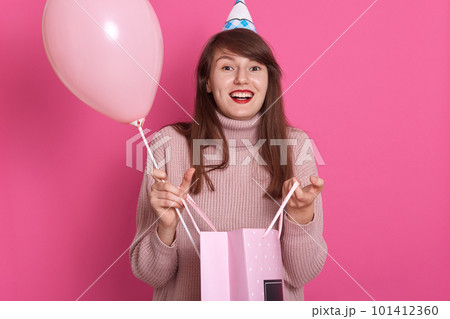 Horizontal shot of young girl with cones on her heads looking directly at camera with happy and surprised facial exprassion, carrying pink balloon and package with gift, isolated over rosy background. 101412360