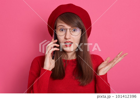 Photo of astonished girl wearing red beret, sweater,spectacles, talking on smartphone with her friend, spreading hand aside, keeps mouth opened, model posing isolated over rose background in studio. Photo of astonished girl wearing red beret, sweater,spectacles, talking on smartphone with her friend, spreading hand aside, keeps mouth opened, model posing isolated over rose background in studio. 101412396