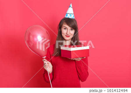 Indoor picture of magnetic lovely young woman having holiday cap on head, holding red baloon and red and white present box, looking directly at camera, wearing bright red sweater. Holiday concept. 101412494