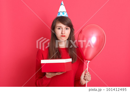Studio shot of young woman girl posing isolated over red background. Valentine's Day, birthday concept, female holding air balloons and present box, looks upset and sad, lady wears beret and sweater. 101412495