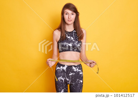 Photo of slim lady wearing sports clothing, holding measure tape, measuring her wrist, comparing her results after fitness exercises, posing against yellow studio wall. Sport and keeping fit concept. 101412517