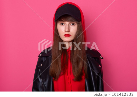 Horizontal shot of young beautiful fashionable teenager girl wearing leather jacket, red hoody and black cap, looking directly at camera, looks confident and serious, model posing with red lips. Horizontal shot of young beautiful fashionable teenager girl wearing leather jacket, red hoody and black cap, looking directly at camera, looks confident and serious, model posing with red lips. 101412535