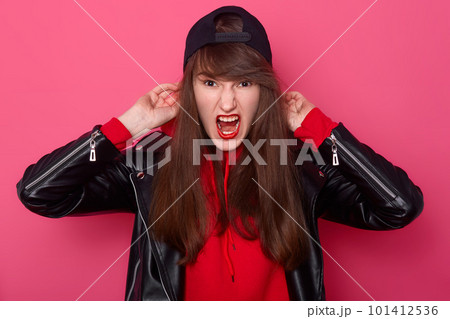 Front view portrait of angry girl with long hair and makeup in casual style black leather jacket standing and screaming in studio, isolated on pink background, has bad spirit and angry expression. Front view portrait of angry girl with long hair and makeup in casual style black leather jacket standing and screaming in studio, isolated on pink background, has bad spirit and angry expression. 101412536