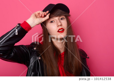 Indoor shot of happy Caucasian young teenager girl in red hoodie, leather jacket, stands with cheeky facial expression, keeps hand on black visor of cap against rose studio wall. People concept Indoor shot of happy Caucasian young teenager girl in red hoodie, leather jacket, stands with cheeky facial expression, keeps hand on black visor of cap against rose studio wall. People concept 101412538