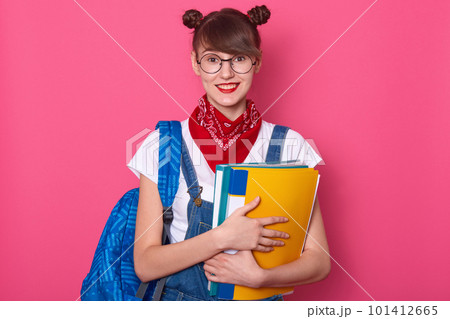 Horizontal shot of peasant looking woman with two hair buns, has delighted expression, holding blue and yellow folder, dresses casual clothing, posing after having classes, isolated over pink wall. Horizontal shot of peasant looking woman with two hair buns, has delighted expression, holding blue and yellow folder, dresses casual clothing, posing after having classes, isolated over pink wall. 101412665