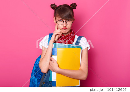 Horizontal view of thoughtful schoolgirl purses lips, keeps index finger on chin, wears round glasses, bandanas on neck, white t shirt, denim overalls, thinks about homework, stands over pink wall. 101412695