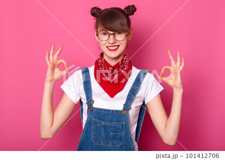 Brunette student with banches smiles, makes okay sign with both hands, shows approval. Young girl wears white t shirt, denim overalls with red bandana on neck. Body language and people concept. 101412706