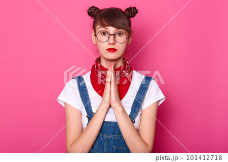 Portrait of black haired brown eyed young female standing isolated over pink background, praying with putting hands together, looking directly at camera, having bunches and bright colored lips. 101412718