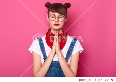 Photo of calm beautiful Caucasian female keeps eyes closed, prays while standing against pink wall, asks God for something important, wears white t shirt, denim overalls, bandana on neck and glasses. Photo of calm beautiful Caucasian female keeps eyes closed, prays while standing against pink wall, asks God for something important, wears white t shirt, denim overalls, bandana on neck and glasses. 101412719