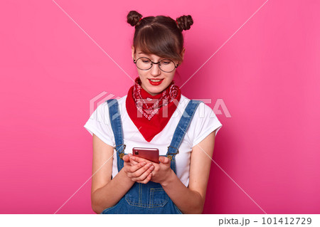 Surprised satisfied cute female typing messages on her phone, watching videos, having bunches on her head, wearing denim stylish overall, white t shirt, red bandana and round stylish eyeglasses. Surprised satisfied cute female typing messages on her phone, watching videos, having bunches on her head, wearing denim stylish overall, white t shirt, red bandana and round stylish eyeglasses. 101412729