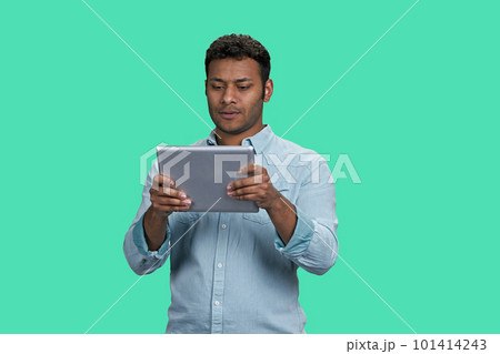 Portrait of a young thoughtful indian guy holding tablet pc. South asian man readin gsomething on his device. Isolated on green background. 101414243