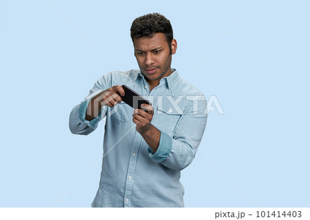 Excited young south asian man playing game in mobile videogame. Isolated on pastel blue background. Excited young south asian man playing game in mobile videogame. Isolated on pastel blue background. 101414403