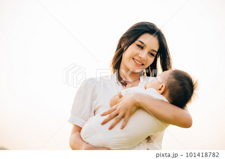 In a quiet park at dusk, a mother holds her sleeping baby, feeling warmth of setting sun. Her face is full of love and contentment as she takes in the beauty of garden. A perfect Mother's Day moment In a quiet park at dusk, a mother holds her sleeping baby, feeling warmth of setting sun. Her face is full of love and contentment as she takes in the beauty of garden. A perfect Mother's Day moment 101418782